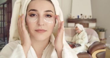 Beyaz bornoz ve göz jeli bandı kullanan beyaz saçlı genç beyaz bir kadının yakın çekimi. Arka plandaki dergide oturan bulanık elbiseli kız. Sinema 4k görüntüleri Prores Hq