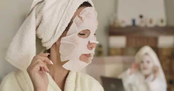 Adult Caucasian woman in hair towel waving to her friend sitting at the background and turning at camera. Pretty girls with face masks resting indoors. Cinema 4k footage ProRes HQ.. — Stock Video