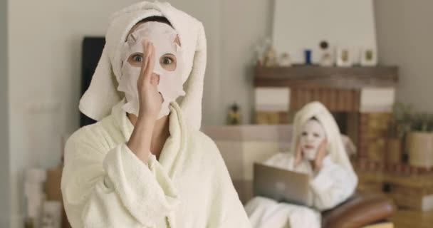 Retrato de mulher caucasiana alegre com máscara facial olhando para trás em amigo ou irmã, e fazendo rostos na câmera. Mulheres positivas em toalhas de cabelo e roupões de banho em casa. Imagens de Cinema4k ProRes HQ . — Vídeo de Stock