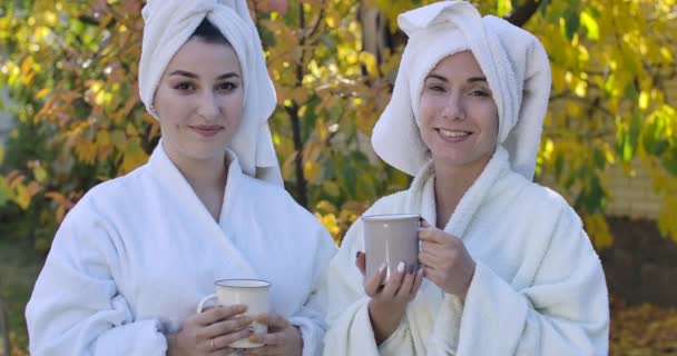 Dos jóvenes mujeres caucásicas lindas en albornoces y toallas de pelo de pie al aire libre, mirando a la cámara y sonriendo. Hermosas chicas descansando en casa. Fiesta de gallinas. Cinema 4k metraje ProRes HQ . — Vídeos de Stock