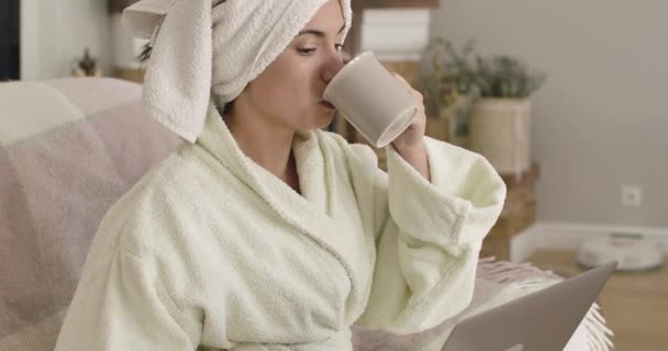 Joven dama caucásica reflexiva en albornoz y toalla de pelo sentado con el ordenador portátil y beber té o café de la taza. Chica bonita usando las redes sociales en casa. Cinema 4k metraje ProRes HQ . — Vídeo de stock