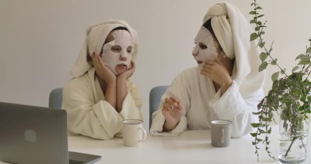 Dwie dorosłe, kaukaskie dziewczyny z maskami na twarzy, siedzące przy stole i gadające. Pozytywne kobiety w ręcznikach do włosów i szlafrokach odpoczywające w domu. Kino 4k materiał Prores Hq. — Wideo stockowe