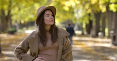 Sonbahar parkının arka planında poz veren zarif kahverengi şapkalı beyaz bir kadın. Neşeli gülümseyen olumlu Avrupalı kız güneşli bir günü dışarıda geçiriyor. Sinema 4k görüntüleri Prores Hq.