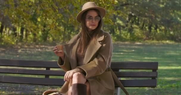 Красивая белая женщина в солнечных очках и элегантной одежде пьет чай или кофе и оглядывается вокруг. Европейская девушка проводит осенний день, сидя на скамейке в парке. Съемки на Cinema4k — стоковое видео