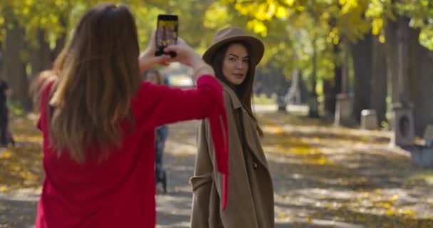 Piękna biała kobieta w eleganckim brązowym płaszczu i kapeluszu, pozująca swojej przyjaciółce na tle jesiennego parku. Pozytywne europejskie kobiety robiące zdjęcia na zewnątrz. Kino 4k materiał Prores Hq. — Wideo stockowe