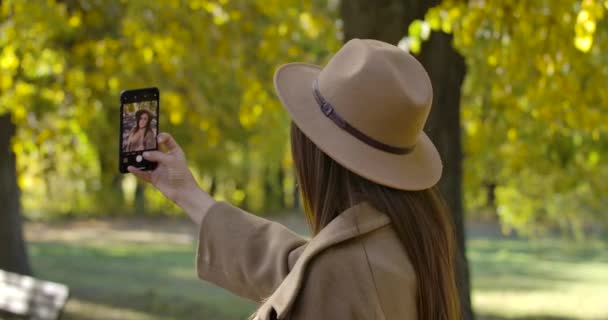 Widok z tyłu młodej białej dziewczyny w brązowym kapeluszu i płaszczu robiącej selfie na smartfonie. Przyjemna Europejka spędzająca weekendy samotnie na świeżym powietrzu. Skupiony na ekranie urządzenia. Kino 4k materiał Prores Hq. — Wideo stockowe