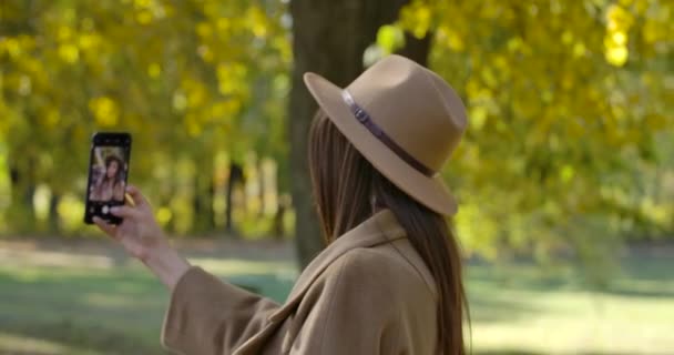 Ung vit flicka i eleganta kläder tar selfie på smart telefon i parken. Trevlig europeisk kvinna tillbringar helger utomhus. Fokusförändringar från förgrund till bakgrund. Film 4k Prores Hq. — Stockvideo