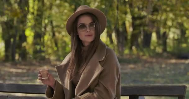 선글라스를 쓰고 갈색 모자를 쓴 매력적 인 백인 여성 이 벤치에 앉아 차나 커피를 마시며 웃고 있습니다. 아름다운 유럽 소녀가 맑은 가을을 밖에서 보내고 있습니다. 영화 4K 필름 프로 레스 HQ — 비디오