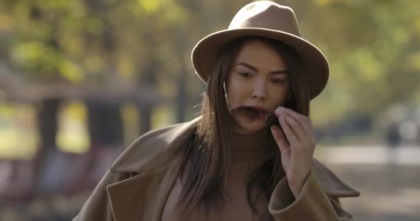 Porträt eines jungen süßen europäischen Mädchens mit elegantem Hut, das seine Sonnenbrille abnimmt und seine langen braunen Haare schüttelt. Charmante Kaukasierin posiert im Herbstpark vor der Kamera. Kino 4k Filmmaterial prores hq. — Stockvideo