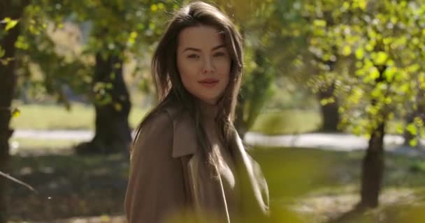 Encantadora chica caucásica con una sonrisa agradable de pie detrás de hojas amarillas en el parque y sacudiendo su largo cabello castaño. Mujer bastante europea posando en la cámara al aire libre. Cinema 4k metraje ProRes HQ . — Vídeo de stock