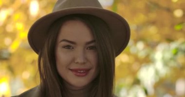 Sarı yaprakların arkasında duran kahverengi şapkalı, çekici, gülümseyen beyaz kadının yakın çekimi. Sonbahar parkında poz veren kendinden emin Avrupalı kız. Sinema 4k görüntüleri Prores Hq.