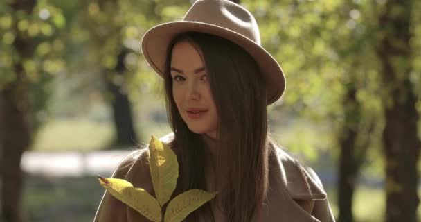 Retrato de menina branca bonito em chapéu marrom no parque de outono olhando para a câmera e sorrindo. Mulher europeia bonita fechando o rosto com folhas, em seguida, movendo ramo de lado. Cinema 4k footage ProRes HQ . — Vídeo de Stock