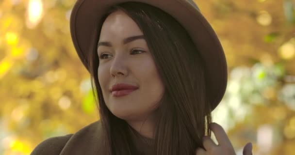 黄色の葉の背景に立って茶色の帽子の魅力的なヨーロッパの女の子のクローズアップ肖像画。公園でポーズをとってカメラに微笑む白人女性を自信満々。映画4k映像｜Prores Hq. — ストック動画