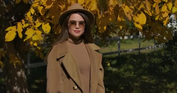 秋の公園のカメラにポーズをとる褐色の帽子とサングラスの若い自信に満ちた白人の女の子。黄色の葉の背景に立つエレガントなヨーロッパの女性。映画4k映像｜Prores Hq. — ストック動画
