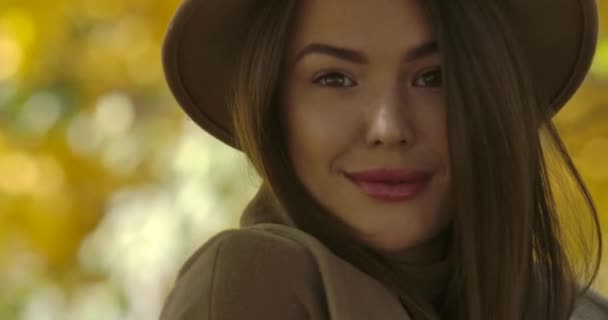 黄色の葉の背景にカメラでポーズ茶色の帽子の若い魅力的なヨーロッパの女性のクローズアップ顔。エレガントな白人の女の子のカメラと笑顔を見て。映画4k映像｜Prores Hq. — ストック動画