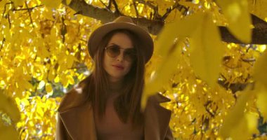 Sonbahar parkında güneş ışığında poz veren güneş gözlüklü, beyaz bir kız. Yüzünde yapraklar olan güzel bir kadın kameraya bakıyor. Sinema 4k görüntüleri Prores Hq.