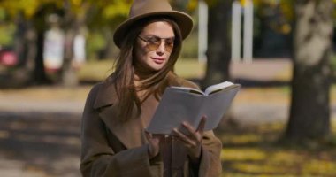 Sonbahar parkında zarif kahverengi elbiseli ve güneş gözlüklü genç beyaz kadın kitap okuyor. Çekici Avrupalı öğrenci hafta sonlarını sonbahar parkında geçiriyor. Sinema 4k görüntüleri Prores Hq.