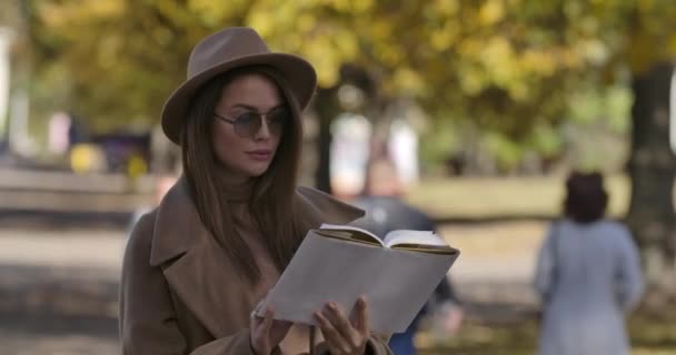 Joven estudiante caucásica en ropa marrón elegante y gafas de sol libro de lectura en el parque de otoño. Encantadora mujer europea inteligente de pie en el parque de otoño. Cinema 4k metraje ProRes HQ . — Vídeos de Stock