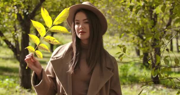 Chica caucásica pensativa en sombrero marrón de pie en el parque de otoño y sosteniendo una rama de árbol. Bonita mujer europea con ojos marrones mirando hacia otro lado y hojas amarillas. Cinema 4k metraje ProRes HQ . — Vídeo de stock