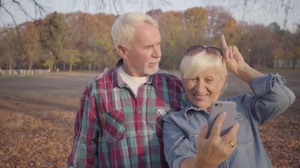 Familia europea senior positiva tomando selfie en el parque de otoño. Alegre pareja moderna caucásica mostrando cuernos y riendo. Envejecer juntos, concepto de amor eterno . — Vídeos de Stock