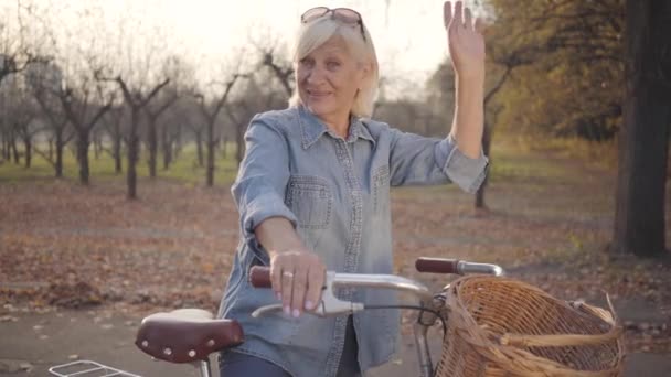 快乐而成熟的白人女士，穿着牛仔，骑着自行车站在那里，对着镜头挥手。 美丽的欧洲退休女人在外面度过秋天的夜晚. 高年级同学假期愉快. — 图库视频影像