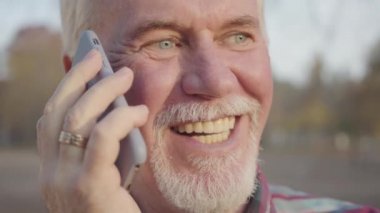 Sonbahar parkında dikilip akıllı telefondan konuşan olgun beyaz adamın yakın plan yüzü. Kıdemli Avrupalı adam konuşmayı bitirip kapatıyor. Yakışıklı emekli erkek, boş zamanın tadını çıkarıyor..