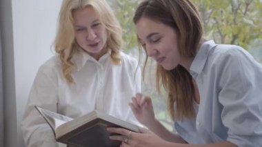 Close-up of young cute Caucasian woman looking photos with older blond lady. Mother and daughter sharing memories at home. Happy family spending leisure time together indoors.
