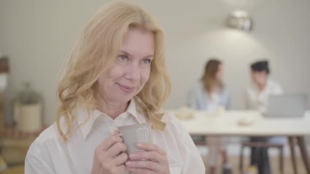 Portrait of beautiful mature Caucasian woman with blond hair and grey eyes. Pleasant senior lady dressed in white shirt standing in the room with a cup of tea or coffee, looking at camera and smiling — Stock Video