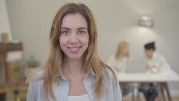 Portrait de jeune belle fille caucasienne aux yeux gris regardant la caméra et souriant. Jolie dame européenne avec un sourire agréable debout dans la chambre sur le fond de deux personnes assises à la table — Video