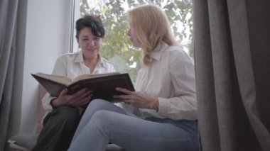 Pencere kenarında oturan iki olgun beyaz kadın fotoğraflara bakıyor. Üst düzey bayan arkadaşlar evde anılarını paylaşıyorlar. Mutlu, gülümseyen hanımlar birlikte vakit geçiriyorlar..