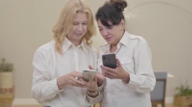 Olgun beyaz kadınlar akıllı telefonla arkadaşlarına öğretiyor. Beyaz tişörtlü iki son sınıf hanımının hafta sonlarını rahat bir evde geçirdikleri portre. Boş zaman, rahatlama, keyif, yeni teknolojiler.