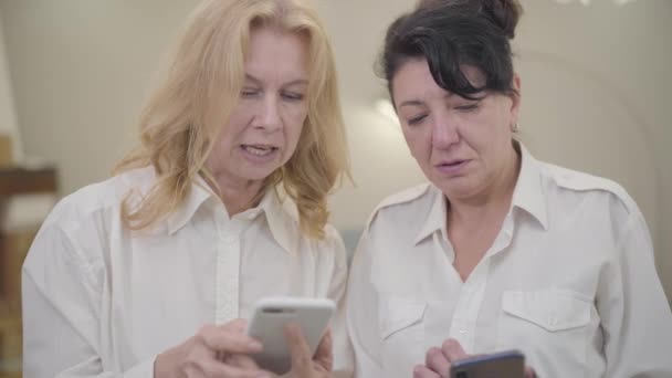 Femmes caucasiennes seniors enseignant à son ami en utilisant le téléphone intelligent. Portrait de deux dames mûres en chemises blanches passant le week-end ensemble à la maison confortable. Loisirs, détente, plaisir, nouvelles technologies . — Video