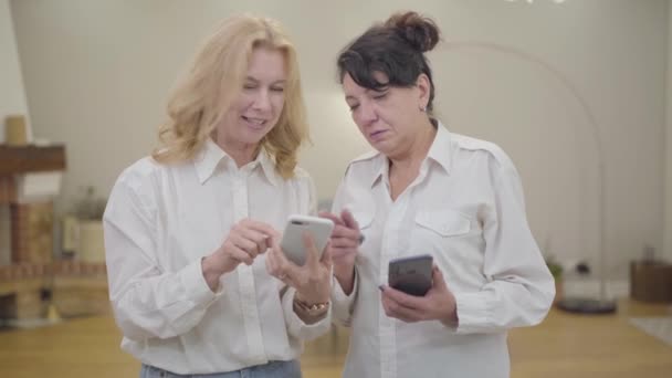 Зрелая белая белая женщина учит своего друга пользоваться смартфоном. Портрет двух пожилых женщин в белых рубашках, проводящих выходные вместе в уютном доме. Отдых, расслабление, удовольствие, новое — стоковое видео