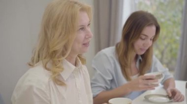 Gülümseyen olgun beyaz kadın misafirlerine komik bir hikaye anlatıyor. Aile, çay içip sohbet etmek için masada toplandı. Genç güzel kız arka planda gülüyor..