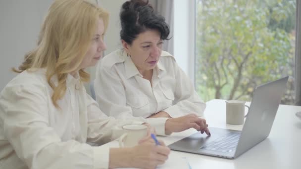 Dos mujeres caucásicas mayores seguras sentadas en la mesa con el ordenador portátil y discutiendo temas de negocios. Mujeres maduras de negocios que trabajan en casa. Nuevas tecnologías en la vida moderna, trabajo freelance . — Vídeo de stock