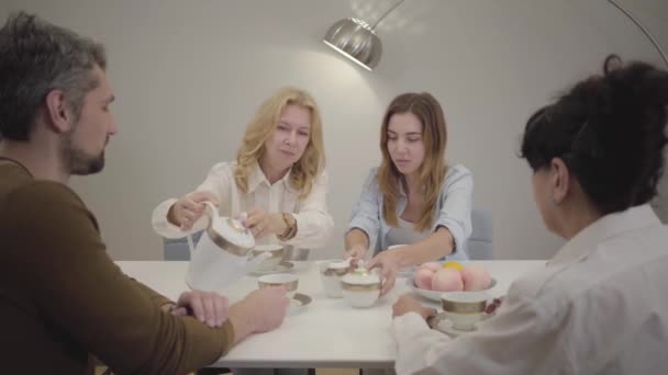 Heureuse famille caucasienne réunie à la table, buvant du thé et bavardant. Femme blonde mature versant une boisson chaude dans des tasses et souriant. Des gens de différents âges célébrant à la maison . — Video