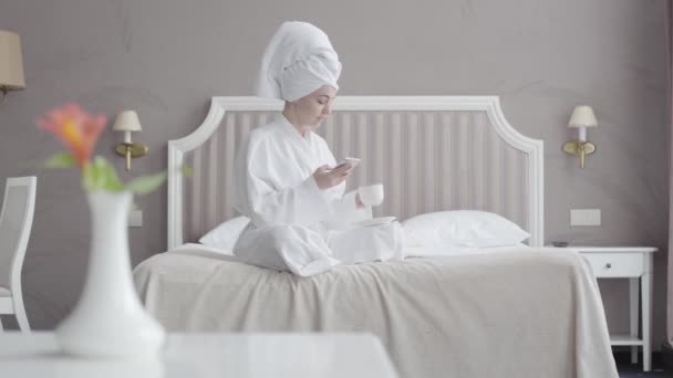 Porträt einer jungen kaukasischen Frau in weißem Bademantel und Haartuch, die morgens Kaffee trinkt und soziale Medien nutzt. Entspanntes Mädchen sitzt auf dem Bett im Hotelzimmer. Tourismus, Freizeit, Lebensstil. — Stockvideo