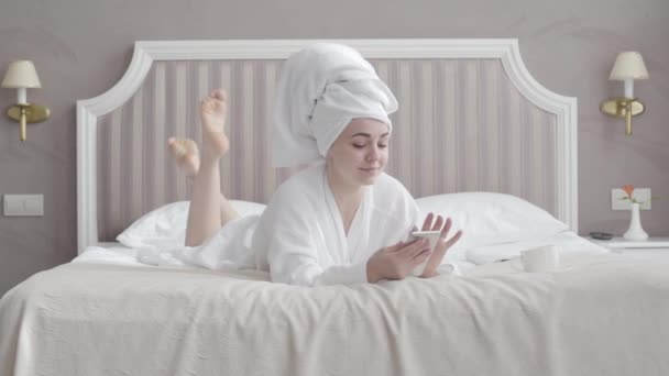 Mujer caucásica relajada positiva en albornoz blanco y toalla de pelo bebiendo café de la mañana y usando teléfono inteligente. Retrato de un joven y hermoso turista acostado en la cama en la habitación de hotel en vacaciones . — Vídeos de Stock