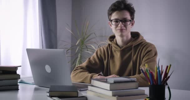 Porträt eines kaukasischen Nerds, der mit Laptop und Büchern am Tisch sitzt und in die Kamera blickt. Lächelnder brünetter Junge mit Brille, der drinnen posiert. Cinema 4k ProRes Hauptquartier. — Stockvideo