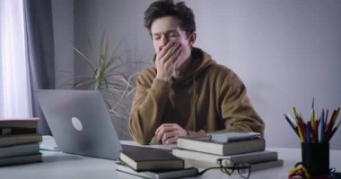 Esmer çocuk esniyor ve pencereden dışarı bakıyor. Ev ödevi yapan çalışkan, beyaz tenli üniversite öğrencisinin portresi. Yaşam tarzı, aşırı çalışma, eğitim. Sinema 4k ProRes Merkezi.