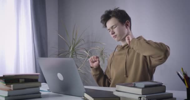 Teenage boy stretching as sitting at the table with laptop and books. Portrait of tired Caucasian college student learning online at home. Education, tiredness, lifestyle. Cinema 4k ProRes HQ. — Stock Video