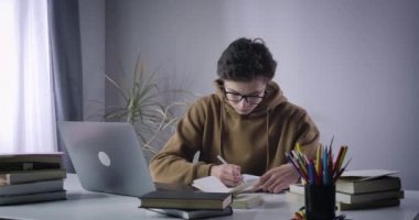 Kafkasyalı çocuk ders kitabını yazıyor ve dizüstü bilgisayar ekranına bakıyor. İnternette çalışan gözlüklü üniversite öğrencisinin portresi. Eğitim konsepti, e-öğrenme. Sinema 4k ProRes Merkezi.