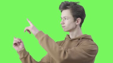 Genç beyaz çocuk parmaklarını kaldırıyor ve ciddi bir yüz ifadesiyle kameraya bakıyor. Gençlik öğrencisi yeşil ekran arkaplanda bir şey gösteriyor. Kromakey.