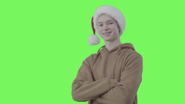 Joyful boy in Christmas hat crossing hands and smiling at camera. Portrait of Caucasian teenager posing at green screen background. New Year, holidays, joy. Chromakey. — Stock Video