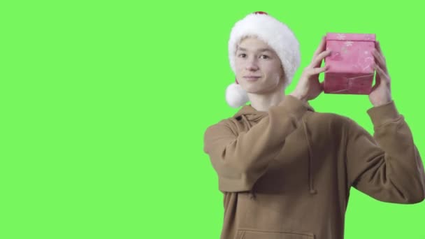 Curious Caucasian millennial boy in New Year hat shaking gift box. Middle shot portrait of smiling teenager receiving present for Christmas. Chromakey, green screen background. — Stock Video