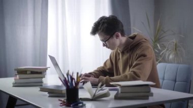 Laptop klavyesinde yazı yazan ve ders kitabında yazan gözlüklü genç dahinin yan görüntüsü. İnternette öğrenim gören Kafkasyalı esmer çocuğun portresi. E-öğrenme, eğitim, zeka.