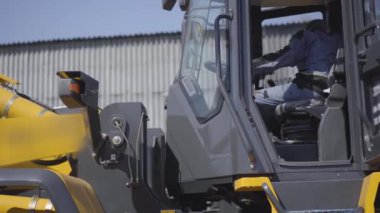 Sarı yükleyicinin kulübesinde oturan Kafkas sürücünün yan görüntüsü. Güneşli bir günde ağır endüstriyel makinelerin içinde üniformalı genç bir çalışan. İş, istihdam, meslek.