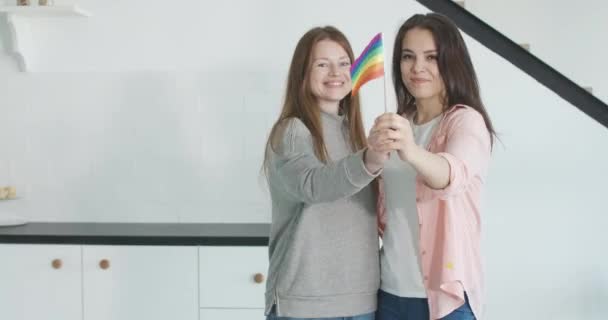 Portrait of happy homosexual couple stretching LGBT rainbow flag to camera. Young cheerful charming confident lesbians. Caucasian same sex family posing indoors. Love concept. Cinema 4k ProRes HQ. — Stock Video