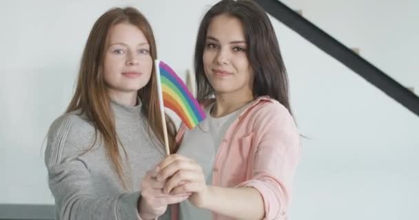 Foco muda de rostos sorridentes de casal lésbico feliz para bandeira do arco-íris LGBT. Retrato do mesmo sexo parceiros do sexo feminino posando com símbolo homossexual dentro de casa. Amor, liberdade, diversidade. Cinema 4k ProRes HQ — Vídeo de Stock