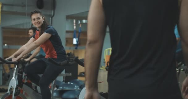 Bonito desportista caucasiano juntar grupo de pessoas atléticas em equipamentos de ciclismo. Jovem positivo cumprimentando homens e mulheres no ginásio andando em bicicletas de exercício. Cinema 4k ProRes HQ . — Vídeo de Stock